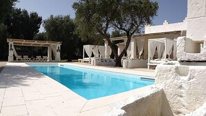 Piscina con suelo de baldosas blancas y una pérgola con cojines blancos.