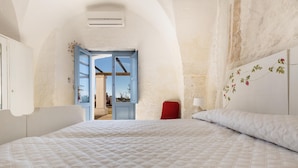Ein Schlafzimmer mit einem großen Bett, einem roten Sessel, einer weißen Wand mit floralem Dekor und einer offenen Tür, die auf einen Balkon führt.