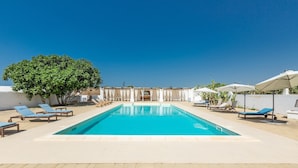 Ein rechteckiger Swimmingpool mit blauem Wasser, umgeben von hell gefliestem Boden. Mehrere Liegestühle mit blauen Kissen sind um den Pool angeordnet.