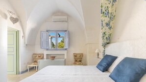 Un dormitorio con una cama grande, una ventana con contraventanas azules, una pequeña mesa con sillas y un tapiz decorativo.