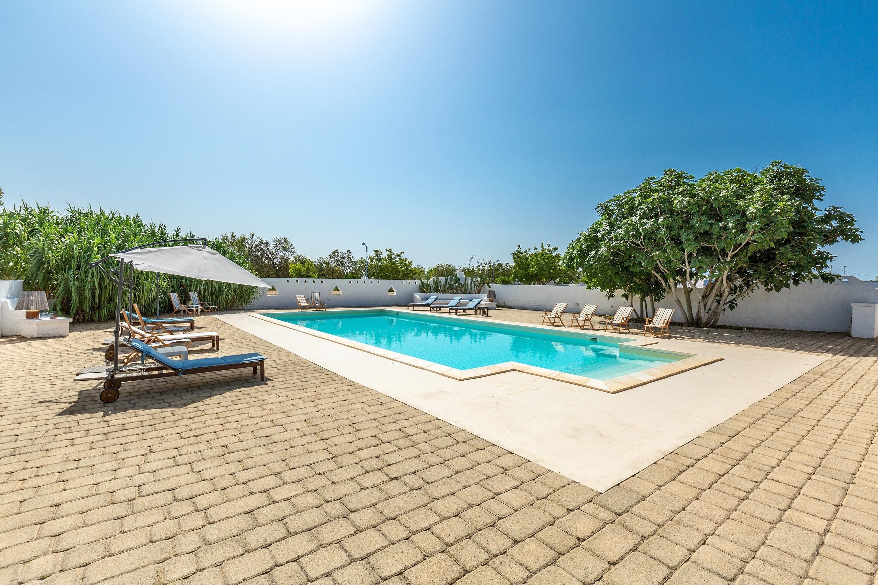 Piscina rectangular con sillas de descanso y una sombrilla, rodeada por un patio empedrado y vegetación.
