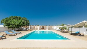 A rectangular swimming pool with blue water, surrounded by light-colored tiled flooring. There are several lounge chairs with blue cushions arranged around the pool.