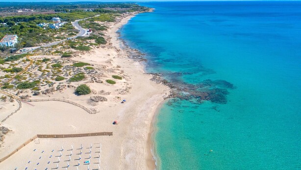 An aerial view of a beach with clear turquoise waters, a sandy shore, and a few umbrellas.