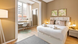 A hotel room with a bed, bedside tables, a desk, and a chair. There are two windows with curtains, a view of buildings outside, and a door leading to another room. - Ola Living Diagonal B 4-2 (Barcelona)