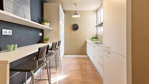 A narrow kitchen with a wooden floor, a white countertop, and a black wall with a clock. There are two pendant lights hanging from the ceiling. - Ola Living Diagonal B 4-2 (Barcelona)