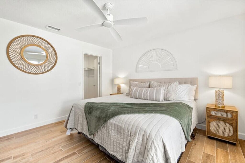 A bedroom with a bed, a ceiling fan, a round mirror, a bedside table with a lamp, and a wooden speaker.
