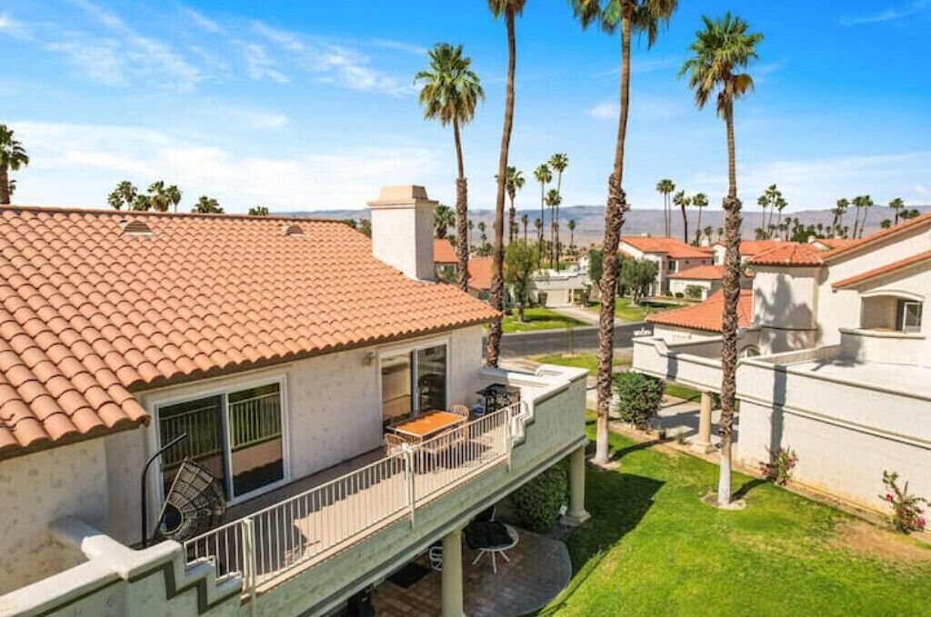 A residential building with a red-tiled roof, a balcony with outdoor furniture, and palm trees in the background.