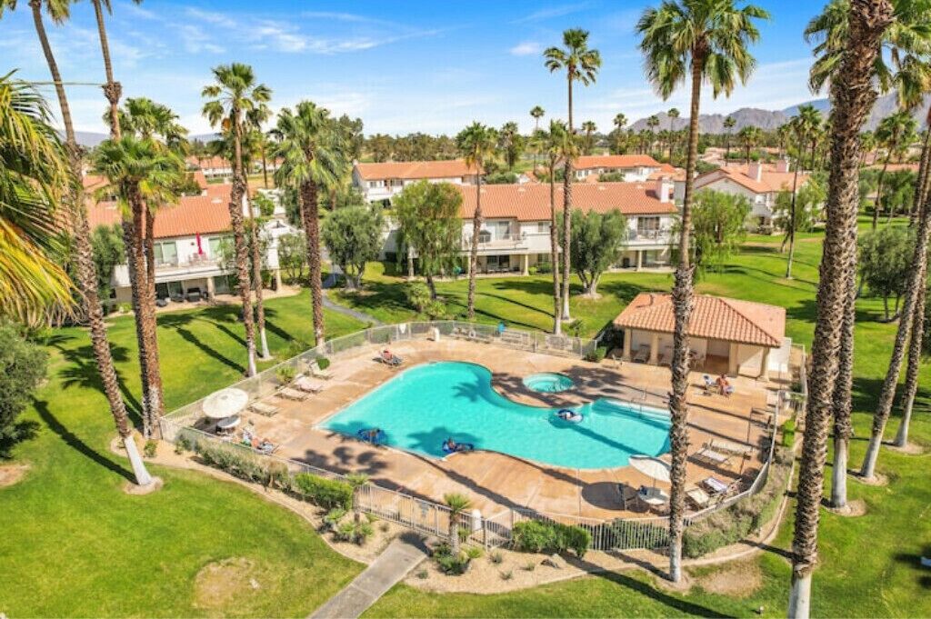 A resort with a large swimming pool, palm trees, and multiple buildings with red-tiled roofs.