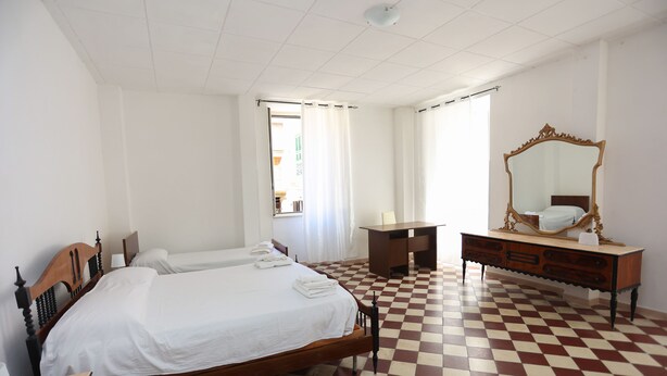 Ett hotellrum med ett rutigt golv, en säng av trä, en byrå med en spegel och ett fönster med gardiner.