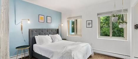 Una camera da letto con un letto, un comodino, una lampada, una finestra vista verde e una pianta pensile.