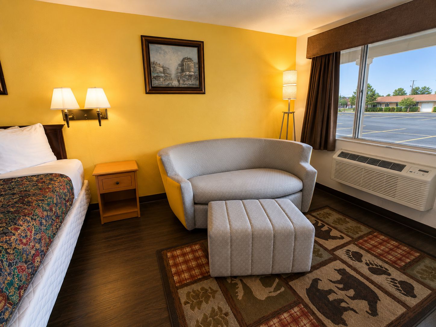 A hotel room with a bed, a gray armchair, a small table, a lamp, a painting, and a window with a view of a parking lot.