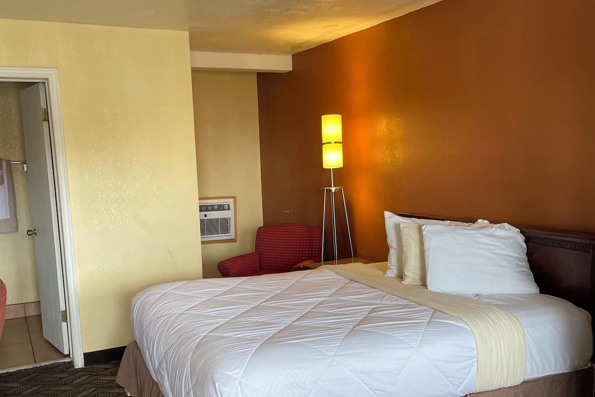 A hotel room with a large bed, a standing lamp, a red chair, and a door leading to another room.