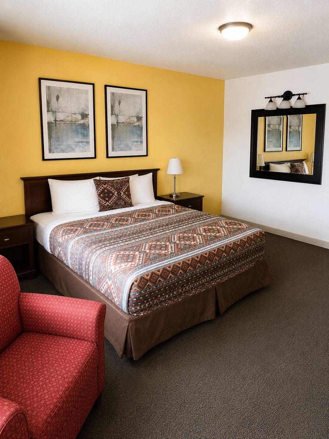 Une chambre d’hôtel avec un lit, une table de chevet, une chaise, une lampe et deux tableaux encadrés au mur.