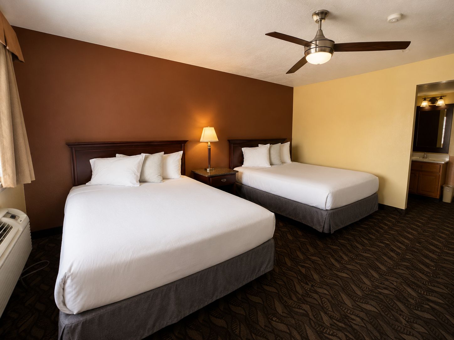 A hotel room with two beds, a ceiling fan, a nightstand with a lamp, and a bathroom visible through an open door.