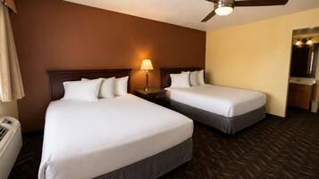 A hotel room with two beds, a ceiling fan, a nightstand with a lamp, and a bathroom visible through an open door.