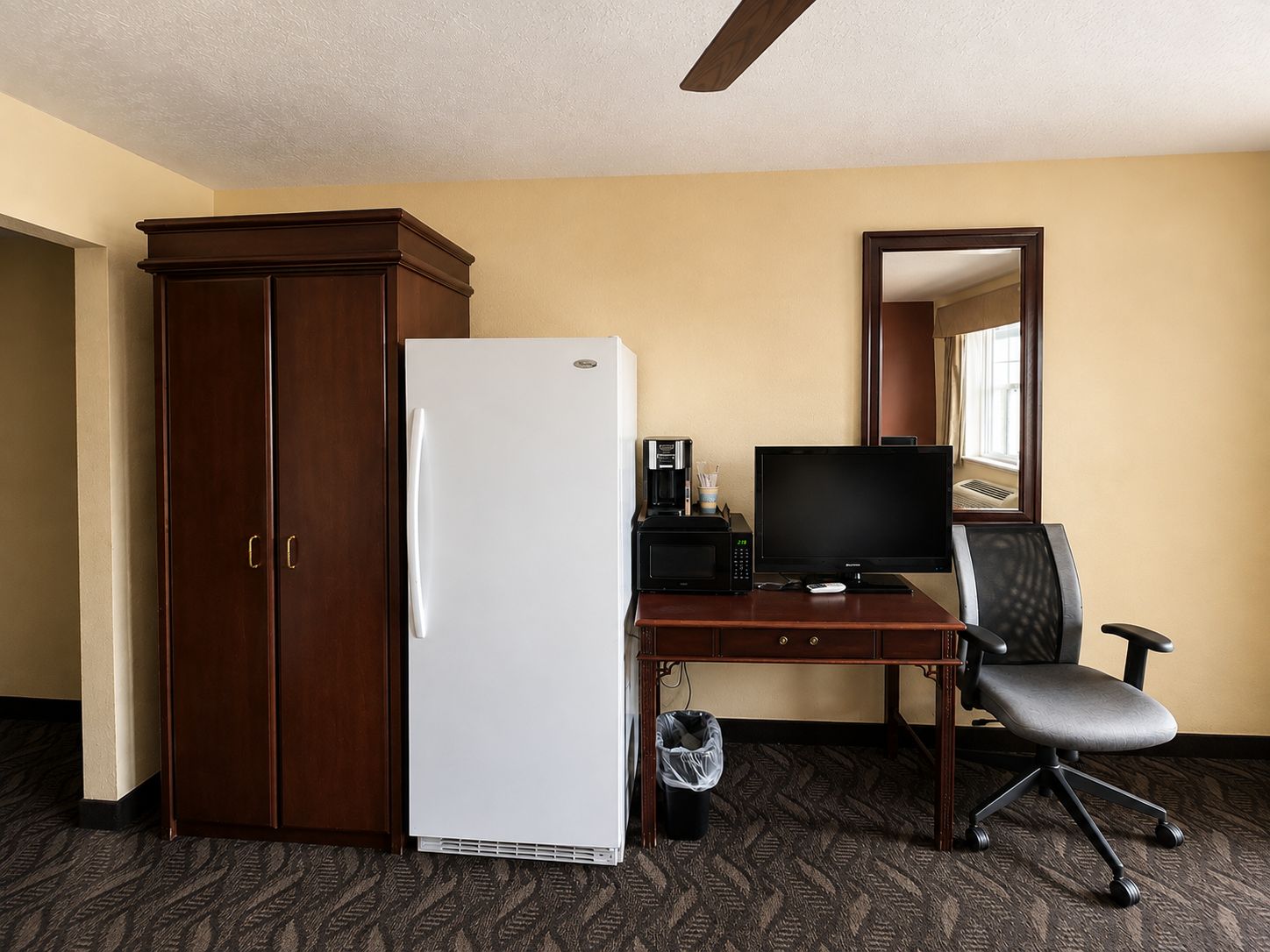 Une chambre d’hôtel avec une armoire en bois, un réfrigérateur blanc, un bureau avec un ordinateur et une chaise, et un miroir au mur.