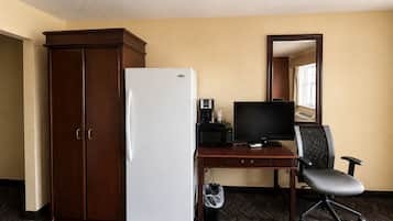 Une chambre d’hôtel avec une armoire en bois, un réfrigérateur blanc, un bureau avec un ordinateur et une chaise, et un miroir au mur.