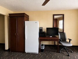 Habitación de hotel con armario de madera, refrigerador blanco, escritorio con computadora y silla, y espejo en la pared.