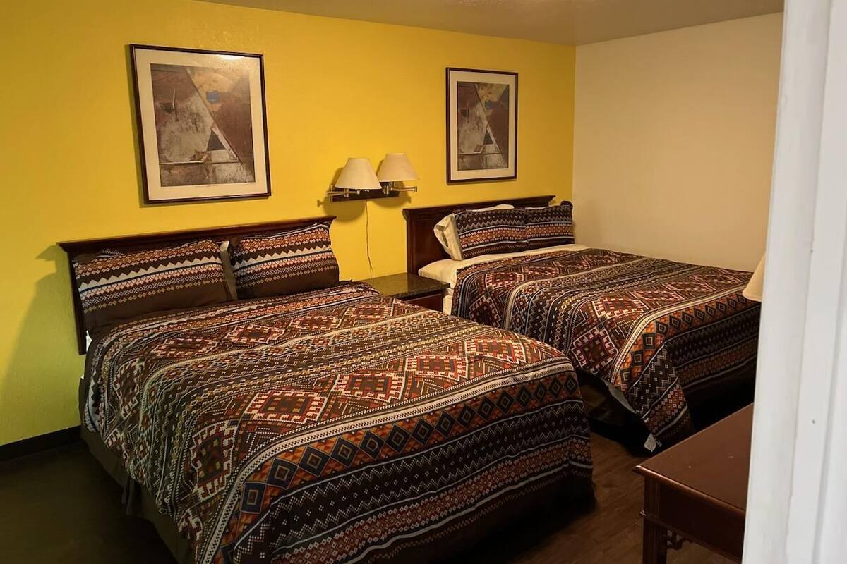 A room with two beds, each with patterned bedding, a nightstand with a lamp, and two framed pictures on the wall.