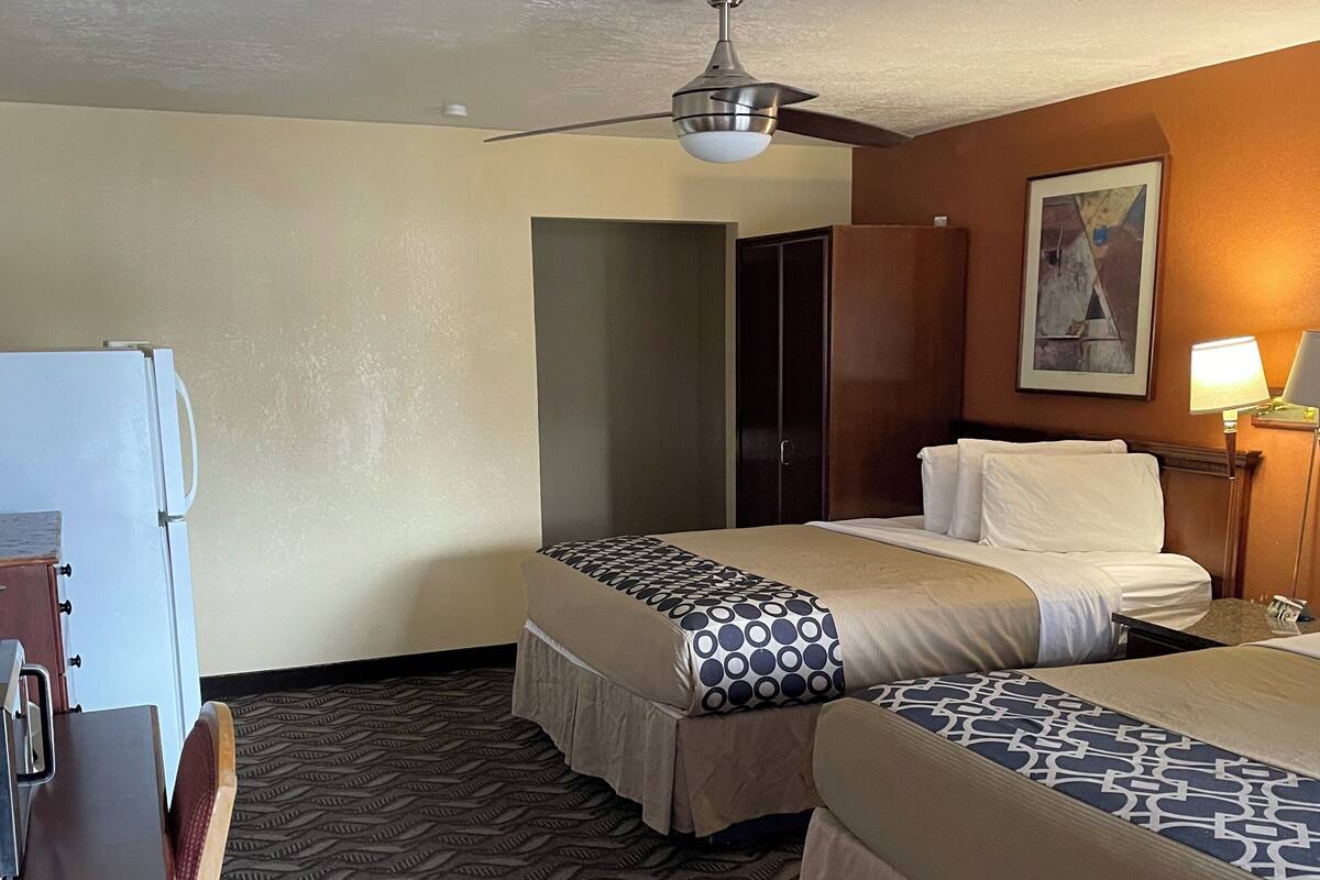A hotel room with two beds, a desk, a refrigerator, a ceiling fan, and a painting on the wall.