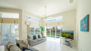 A spacious living room with a large window overlooking a pool, a gray sofa with blue cushions, and a flat-screen TV on a stand. - Stunning Sunsets, Spacious Waterfront Home (Marco Island)