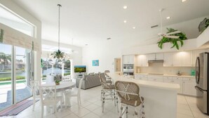 A modern kitchen with white cabinetry, a central island, and a dining area with white chairs. - Stunning Sunsets, Spacious Waterfront Home (Marco Island)