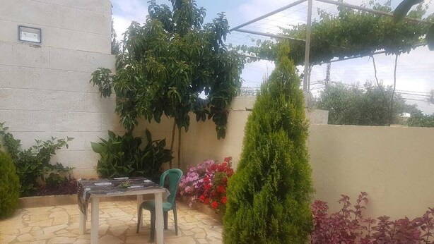 An outdoor patio area with a stone floor, a table with chairs, and various plants and shrubs. - Bright apartment with garden (الكورة)