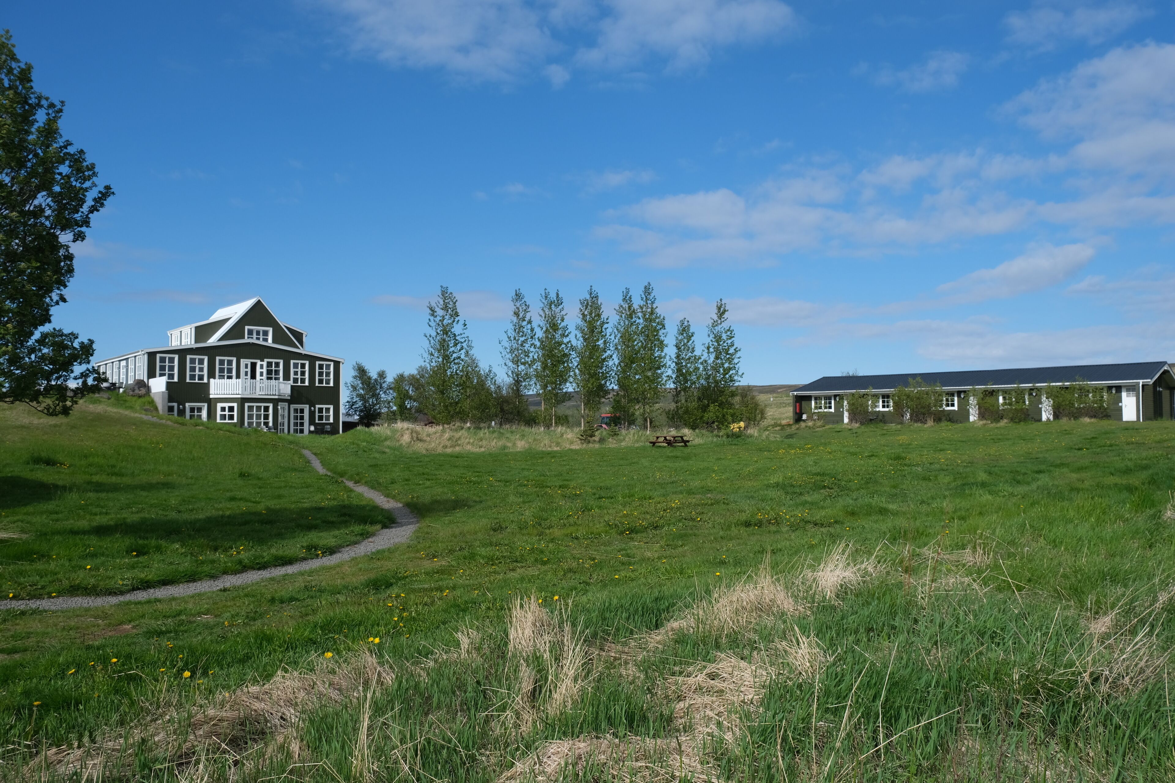A large, multi-story house with a prominent white exterior and a sloped roof, surrounded by a grassy field and trees.