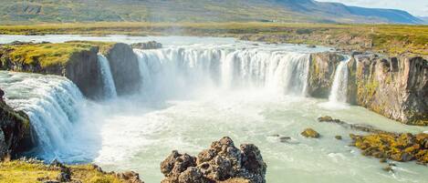 A waterfall cascading over rocky terrain with a river below, surrounded by grassy hills.