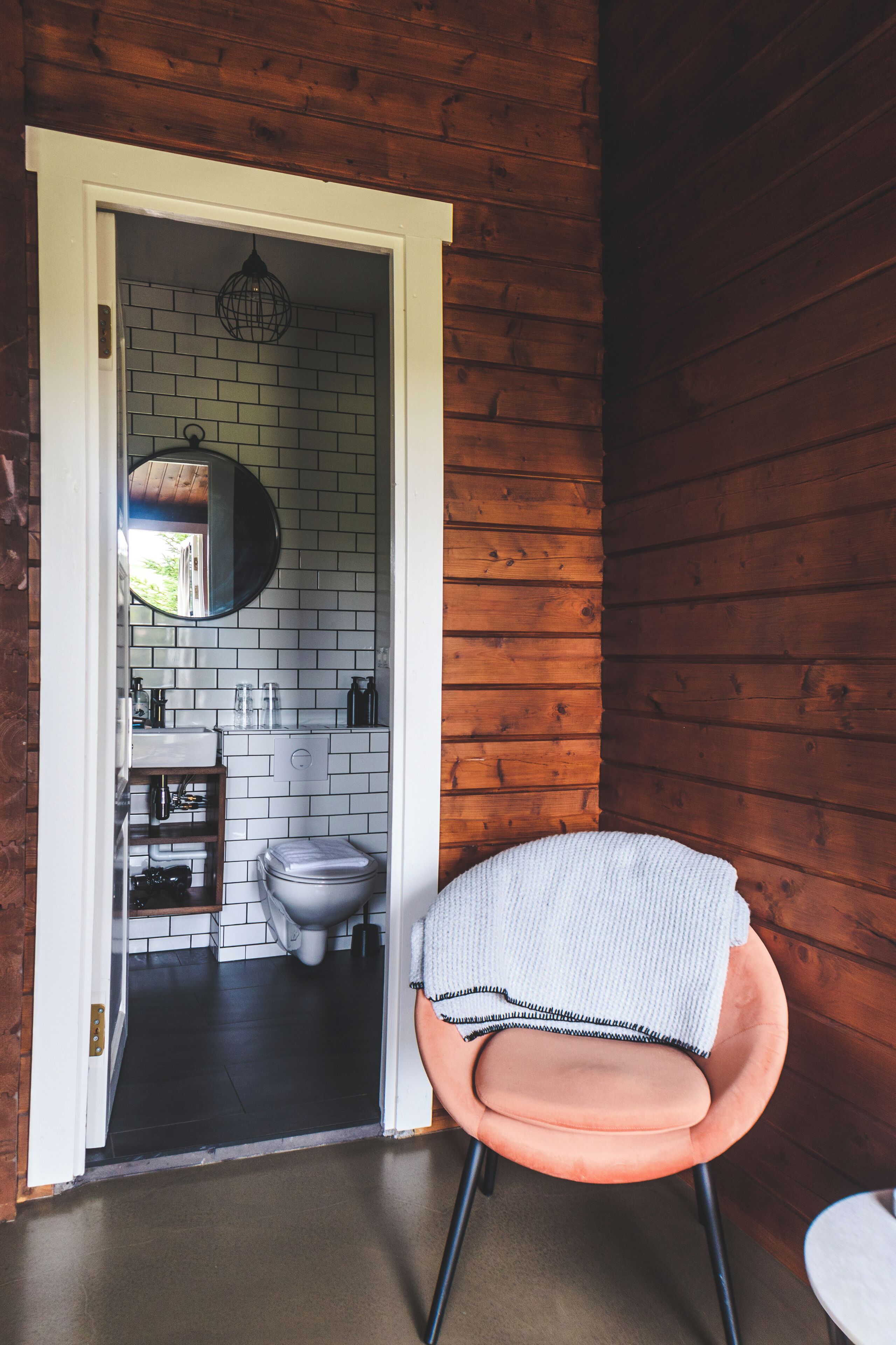 A wooden cabin interior with a white brick wall, a round mirror, a white toilet, and a pink chair.