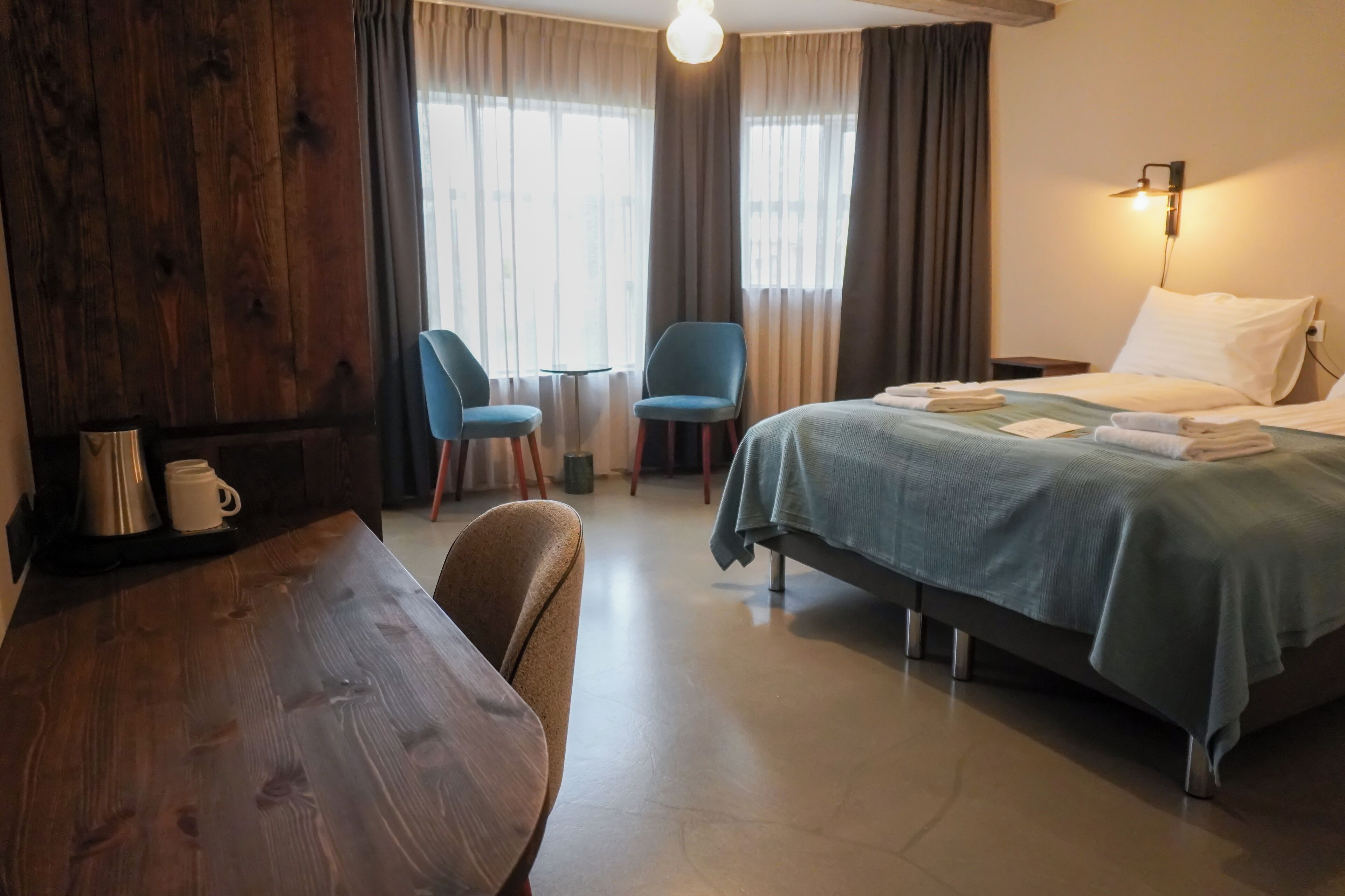 Habitación de hotel con una mesa de madera, dos sillas azules, una cama con colcha azul y una mesita de noche con lámpara.