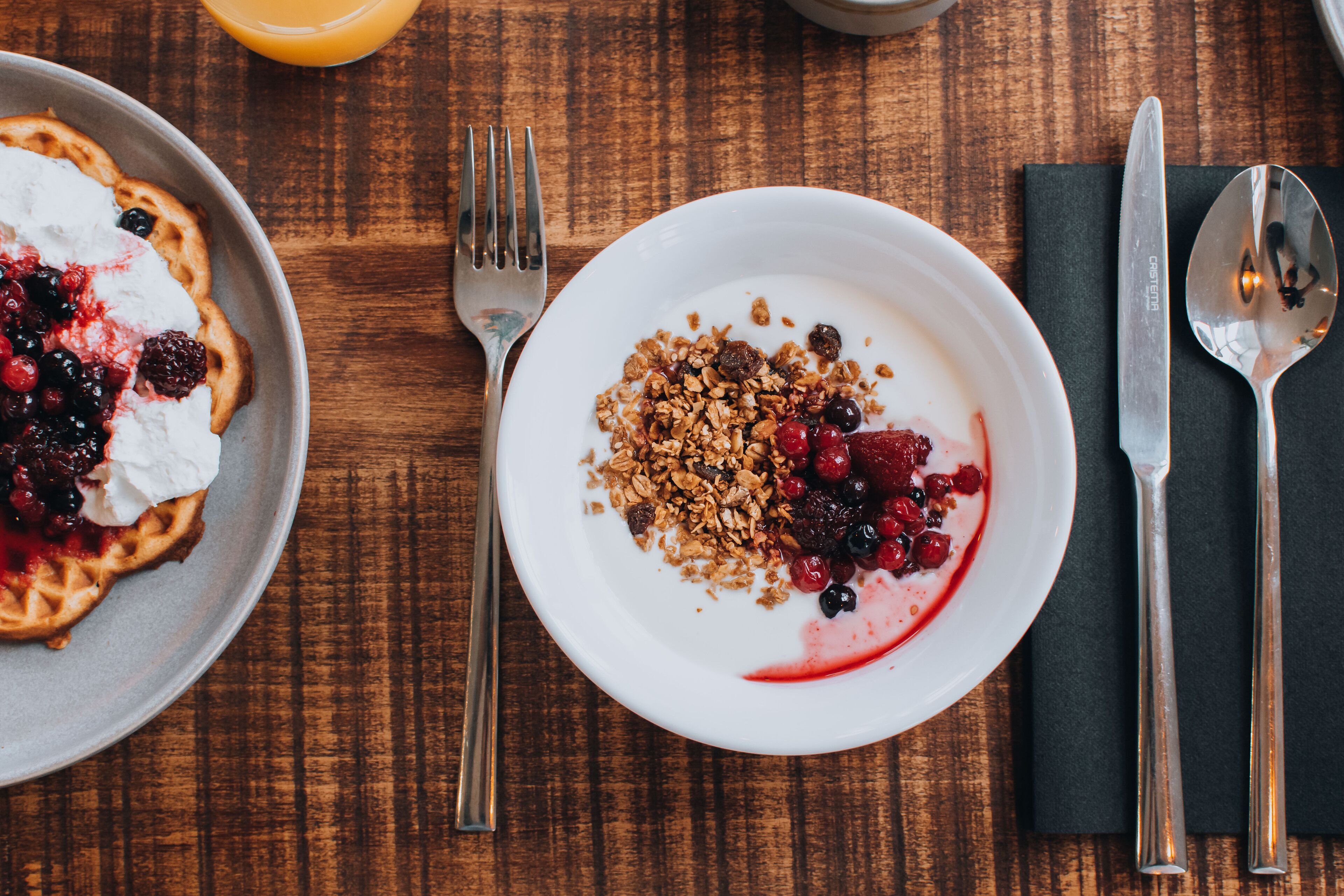 Une table en bois dressée pour le petit-déjeuner, avec des gaufres, du yaourt, du granola et des fruits rouges.