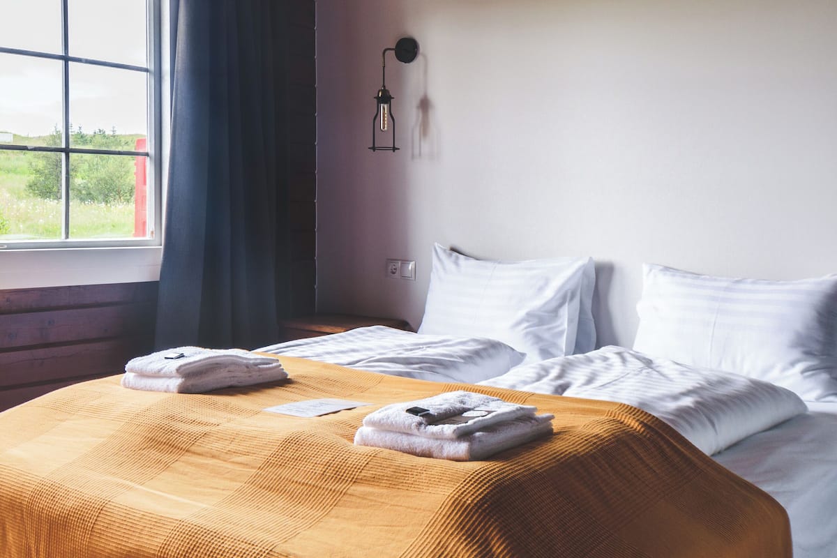 A neatly made bed with white linens, a wooden headboard, and a window with curtains.