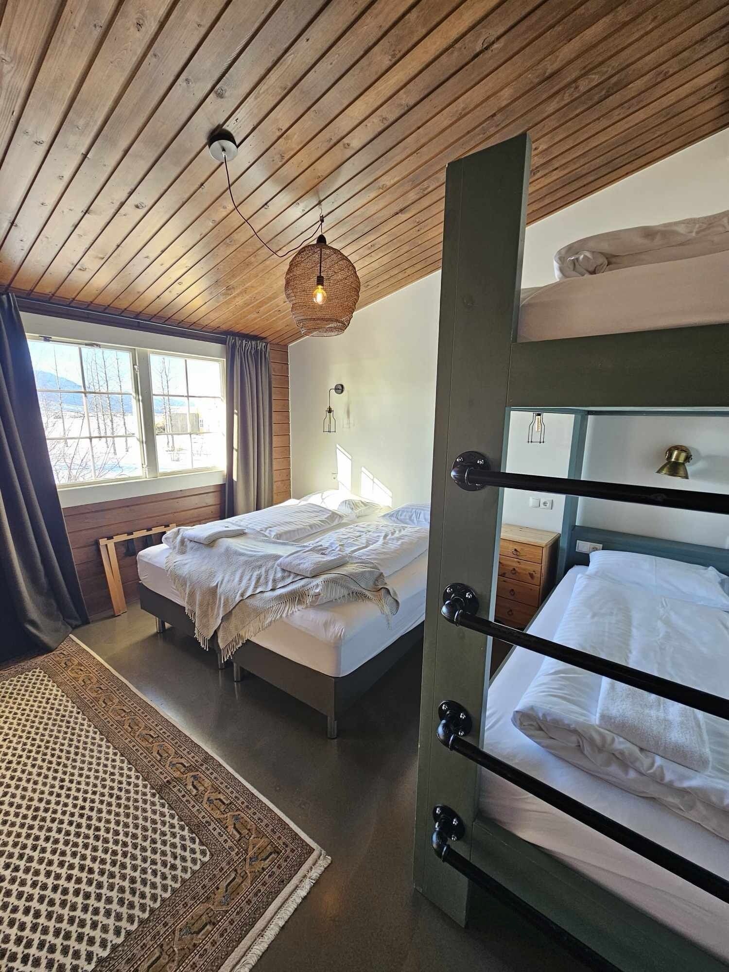 Quarto com duas beliches, teto de madeira, janela grande com vista para montanhas e luminária pendente.