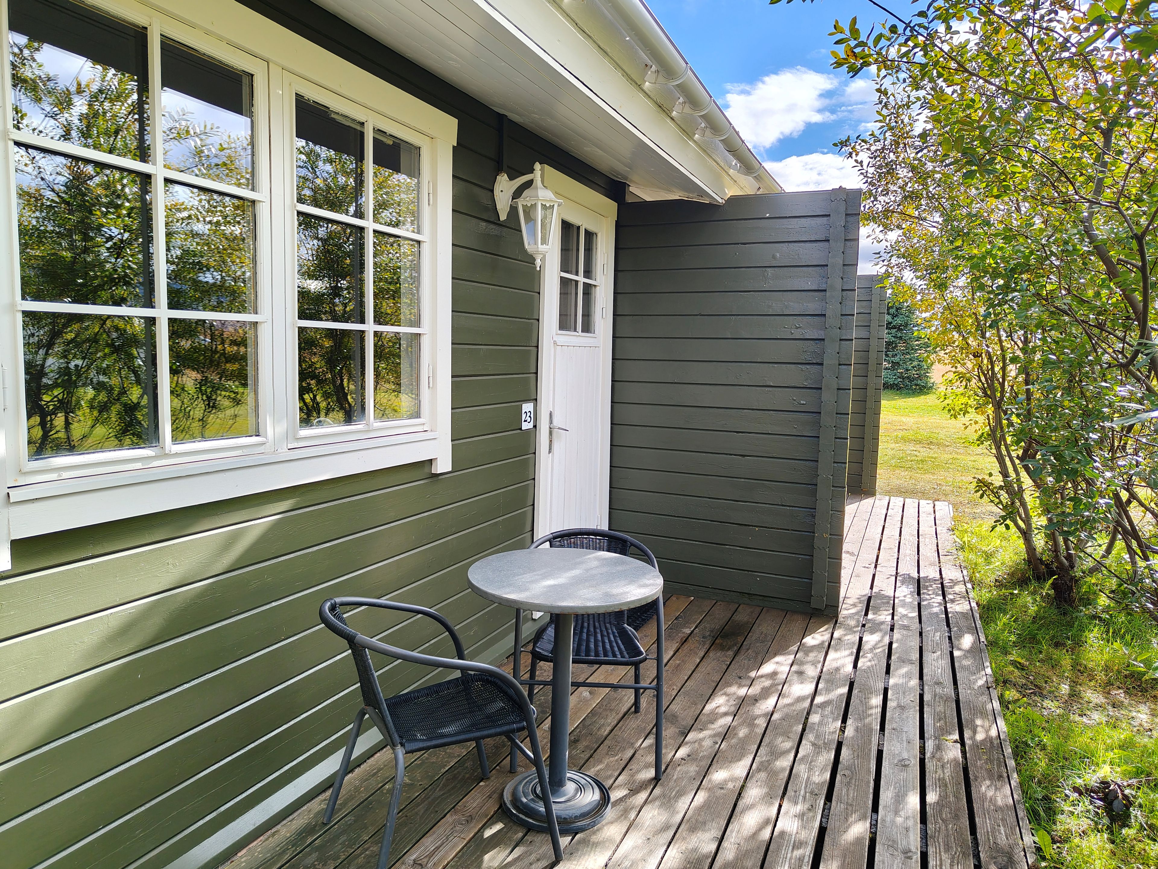 A wooden deck with a table and chairs, a door with a number, and a window with a view of trees.