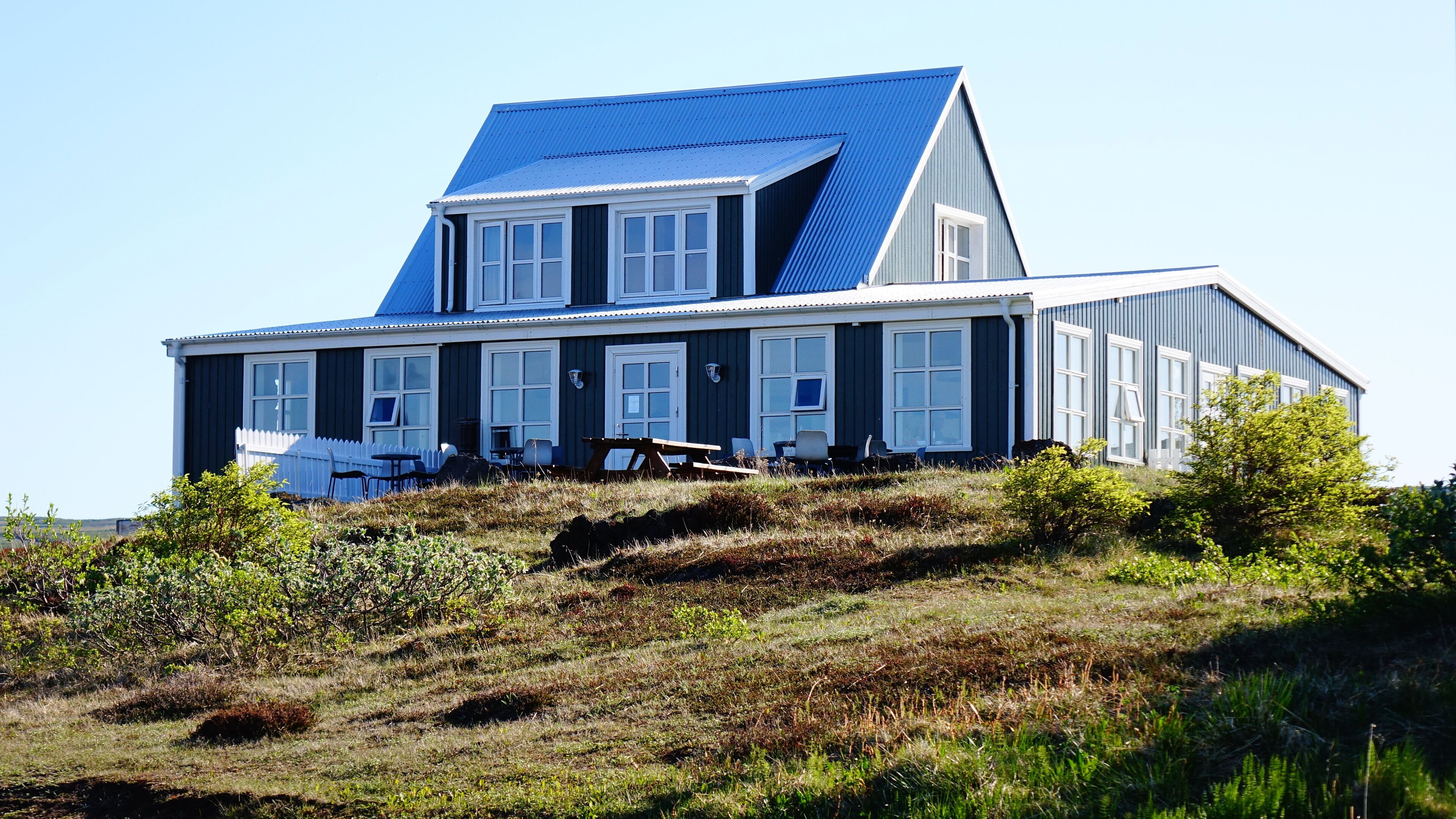 Une maison à deux étages, avec un toit bleu et des boiseries blanches, située sur une colline verdoyante et entourée d’une clôture en bois peinte en blanc.