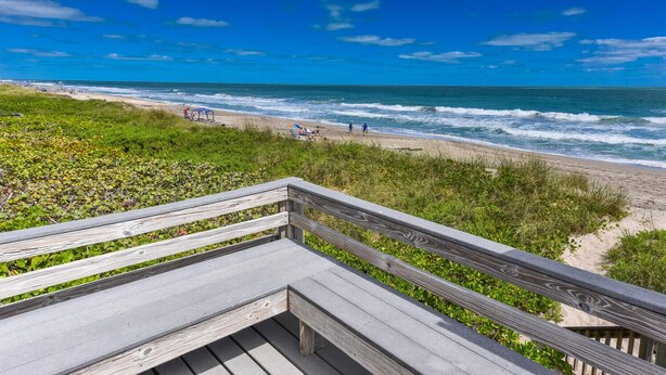 A wooden deck overlooking a beach with waves, grassy areas, and a clear blue sky. - Minute walk to the Beach! Florida Condo with screened in porch. (Stuart)