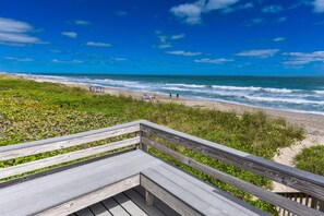 Una terraza de madera con vista a una playa, olas, zonas verdes y un cielo azul despejado.