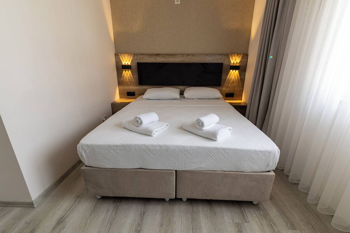 Een netjes opgemaakt bed met twee kussens en twee opgerolde handdoeken, een hoofdbord met een flatscreen televisie en wandlampen voor het lezen.