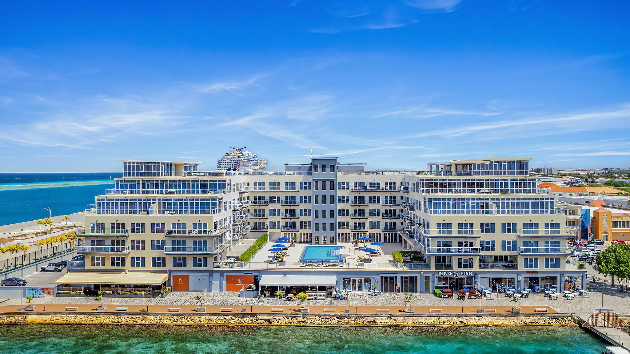 A multi-story hotel with a pool area, located near a body of water.