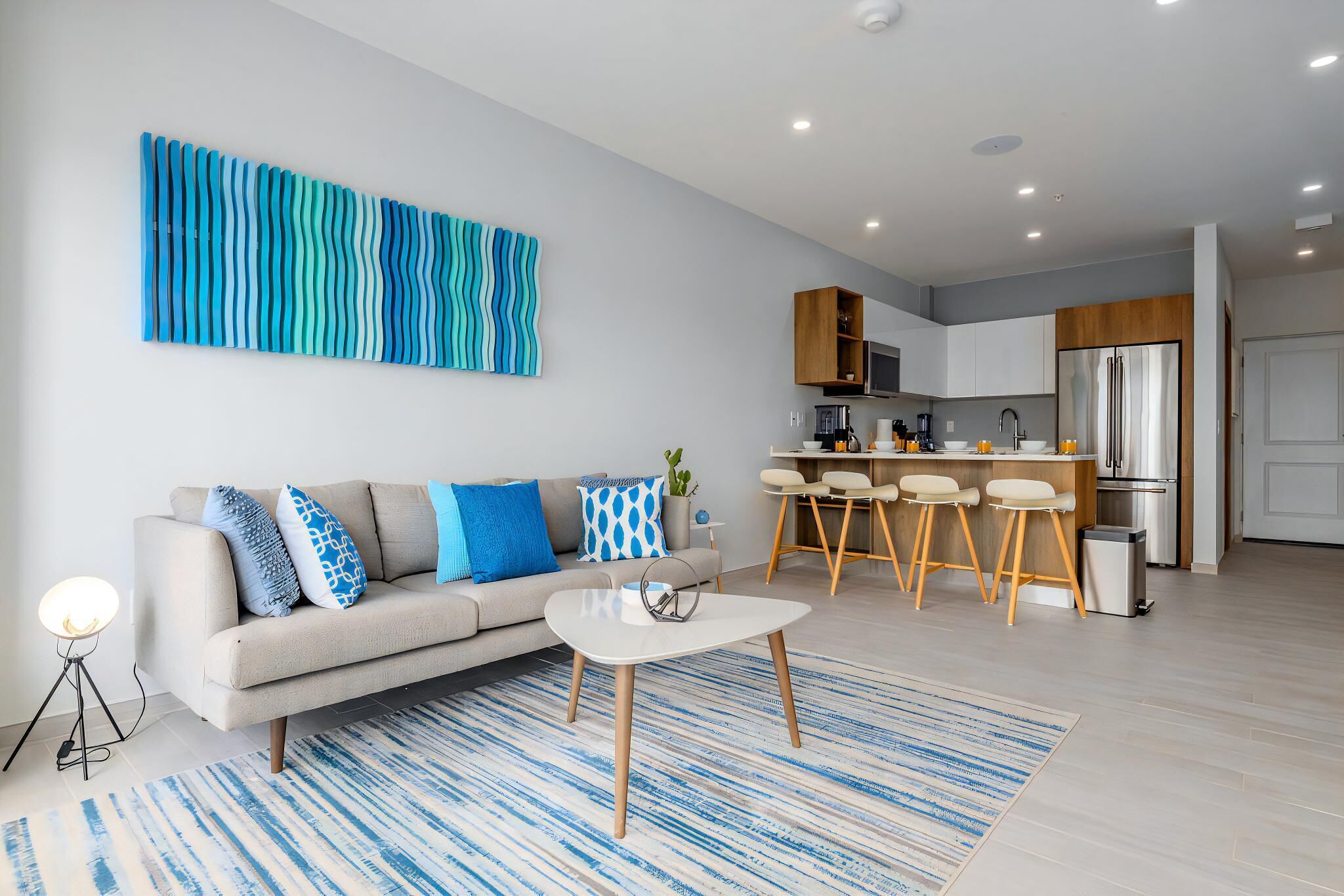 Una sala moderna con un sofá, una mesa de centro y un cuadro abstracto grande. Se ve una cocina con taburetes de bar y un refrigerador al fondo.