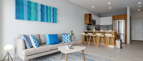 Una sala moderna con un sofá, una mesa de centro y un cuadro abstracto grande. Se ve una cocina con taburetes de bar y un refrigerador al fondo.