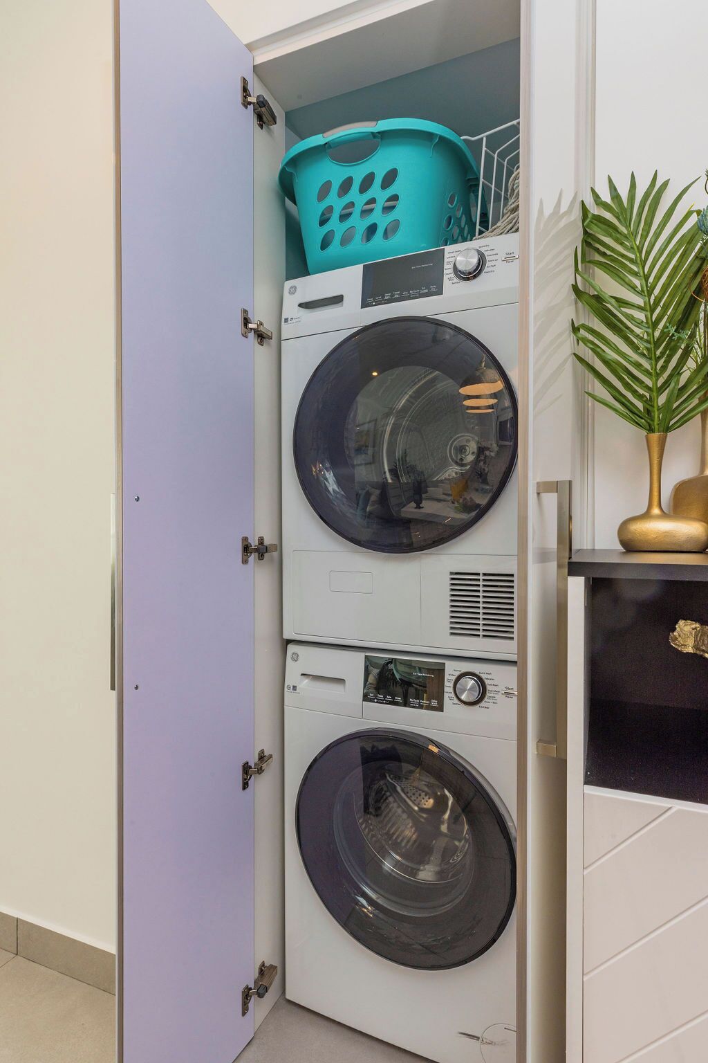 Un cuarto de lavandería con lavadoras y secadoras apiladas, un cesto de ropa de color aguamarina y una planta en maceta.