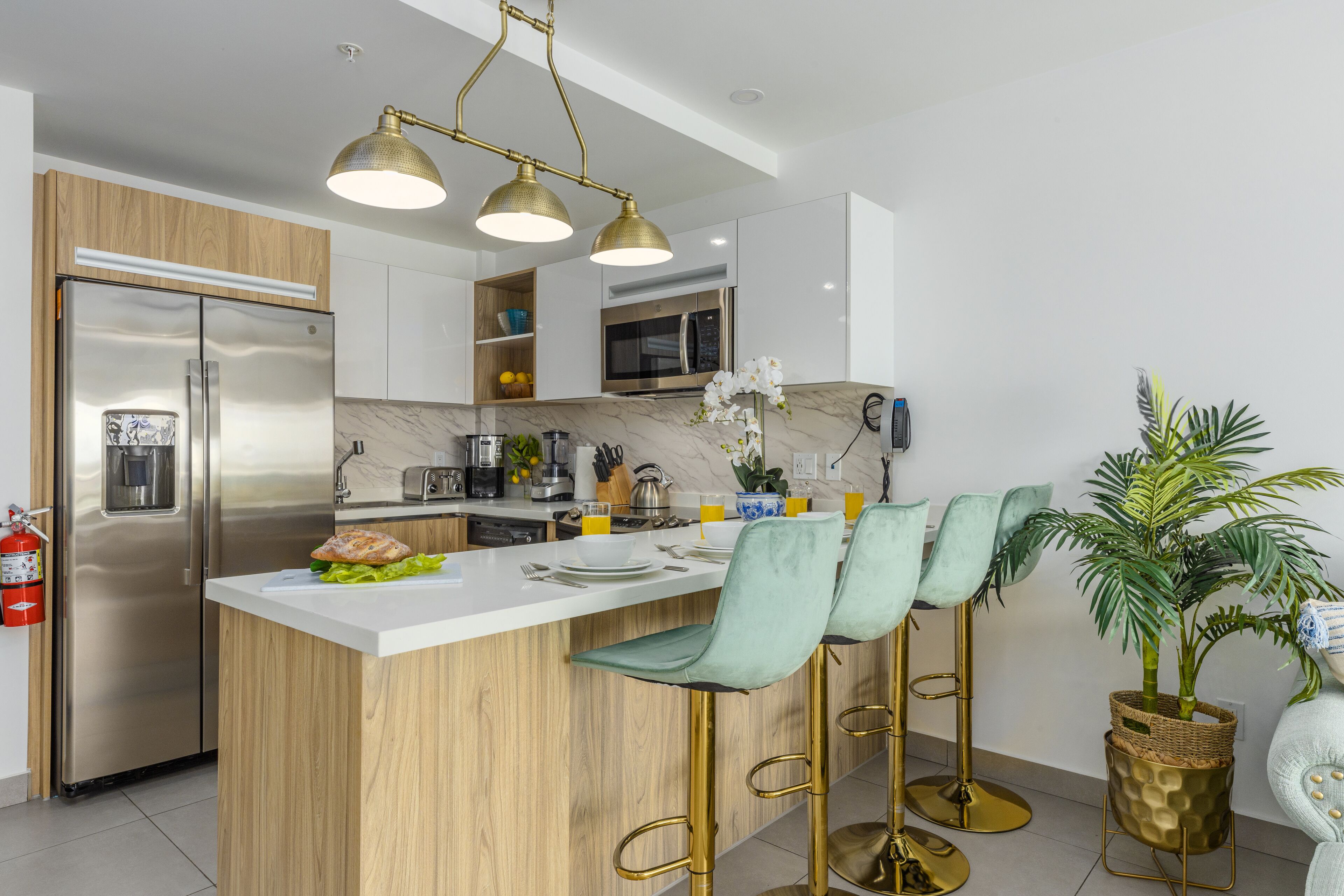 A modern kitchen with a stainless steel refrigerator, wooden cabinets, a marble countertop, and a bar area with green barstools and gold accents.