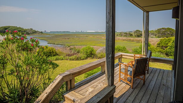 Eine Holzterrasse mit zwei Stühlen und einem Tisch, mit Blick auf eine Wiese mit einem Gewässer und entfernte Gebäude.