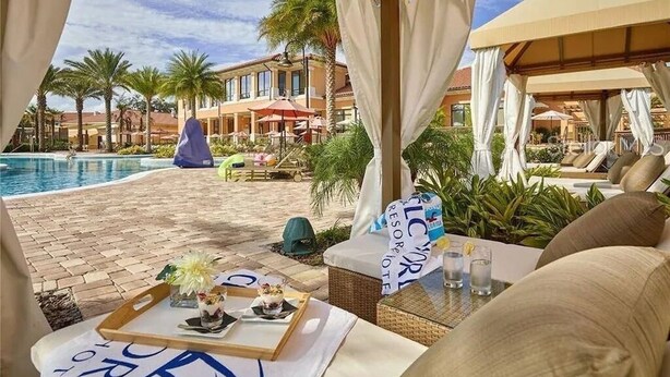 Ein Poolbereich eines Resorts mit überdachter Sitzgelegenheit, Palmen und Blick auf die Hotelgebäude.