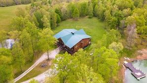 A large log cabin with a green roof surrounded by lush greenery and a dock extending into a lake.
