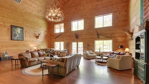 A spacious log cabin interior with multiple seating areas, a fireplace, and large windows allowing natural light.
