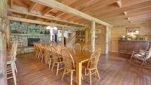 A spacious dining area with a wooden table and chairs, a stone fireplace, and a kitchen area visible in the background.