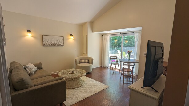 A living room with a sofa, armchair, and a coffee table. There is a television on a stand, a dining table with chairs, and a large window with curtains. - Charming Couple's Cottage close to Lake Michigan, Silver lake, and Stoney Lake (Shelby)